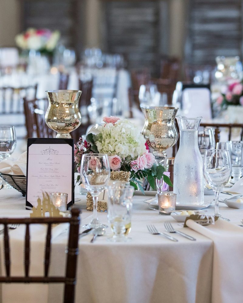 Close-up wedding table detail with menu card, floral centerpiece, and mercury glass vessels